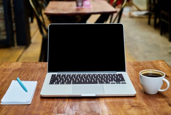 A laptop on a wooden surface, next to a notepad and a cup of coffee.