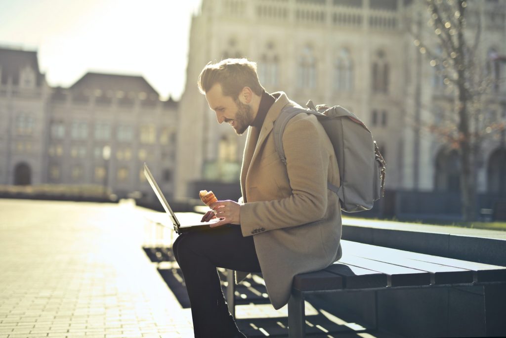 A smiling man using his laptop on a bench in the city while eating a croissant